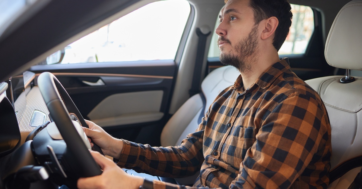 4 Common 6.0L Powerstroke Engine Problems A man wearing a brown-and-black plaid button-down shirt sits behind the wheel of a vehicle, his hands on the wheel.